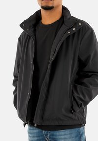 Giacca nera con zip frontale, colletto alto e bottoni a pressione, dotata di tasche laterali e stoffa liscia e leggera. Abbinata a una camicia nera.