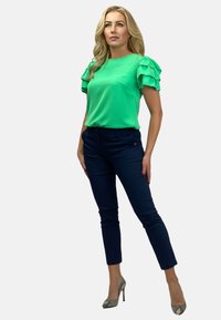 Zielona bluzka z krótkim rękawem i falbaniastymi akcentami, zestawiona z granatowymi dopasowanymi spodniami i szpilkami w wężowy wzór, wystylizowana na neutralnym tle.