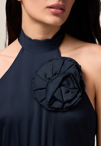Marineblaues Neckholderkleid mit einer großen, strukturierten Blumenapplikation. Glattes, glänzendes Material mit fließendem Design. Schlitzöffnung am Ausschnitt.
