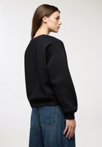 Giovane donna con capelli castano lisci, che indossa un ampio felpa nera e jeans blu, in piedi di profilo contro uno sfondo bianco semplice.