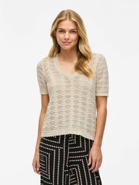 Maglione lavorato a maglia beige chiaro con scollatura a V, maniche corte e un motivo a onde. Indossato sopra una gonna nera con accenti bianchi geometrici.