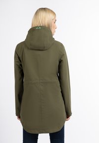 Chaqueta impermeable verde oliva con capucha, detalles en la costura trasera y dobladillo ligeramente curvado. Textura suave y diseño funcional.