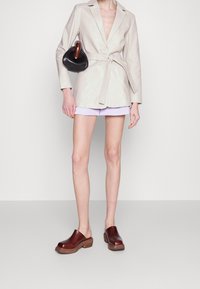 Blazer en cuir beige avec ceinture à la taille, accompagné d'un short lavande. Sabots marron à talon épais, sac à main noir avec une anse en bois.