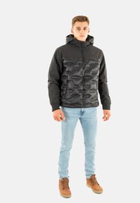 Chaqueta con capucha negra que presenta un diseño frontal acolchado y mangas lisas. El modelo lleva puestos unos pantalones vaqueros azul claro y botas de cordones marrones.