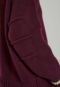 Burgundi színű kötött pulóver, textúrált, bordázott mintázattal. Az ujjon finom varrott részletek láthatók, és a mandzsetta passzos.