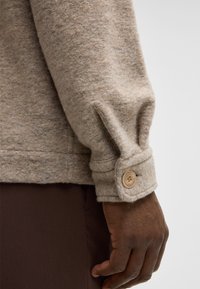 Maglione di lana beige con una finitura testurizzata, dotato di un dettaglio con polsini abbottonati, abbinato a pantaloni marrone scuro, che mette in risalto il calore del tessuto.