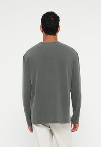 Mann mit kurzen, lockigen schwarzen Haaren, der ein langärmliges, dunkelgraues Hemd und hellbeige Hosen trägt, von hinten vor einem schlichten weißen Hintergrund betrachtet.