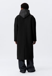 Persona con un abrigo largo negro con capucha gris oscuro adjunta, de pie de espaldas, combinando con pantalones sueltos oscuros y zapatos negros.