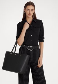 Borsa tote in pelle nera con finitura testurizzata e accenti dorati, tenuta da una persona che indossa una maglietta nera e pantaloni a righe.