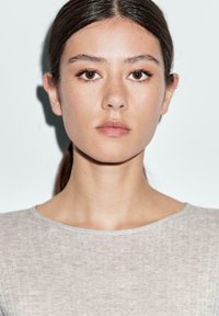 Young woman with brown eyes and hair pulled back, wearing a beige textured top, looking directly at the camera with a neutral expression.