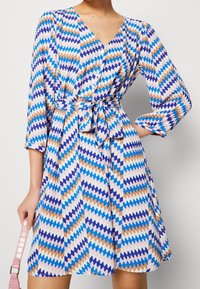 Musterkleid mit geometrischem Zickzackdesign in Blau, Orange und Weiß. V-Ausschnitt, lange Ärmel und Taillenschnürung. Weicher Stoff.