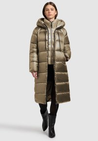 Gewatteerde lange jas in metallic olijfgroen. Voorzien van een capuchon, ritssluiting en zwarte accenten. Soepele textuur met een doorgestikt patroon.