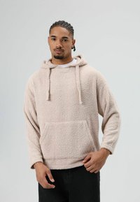 Beige, pluizige hoodie met een voorzak, trekkoorden en lange mouwen. Zachte textuur, casual ontwerp. Gedragen over een wit T-shirt en zwarte broek.