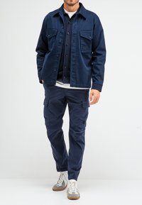 Marineblaues Freizeitjackett mit zwei Brusttaschen, über einem Hemd getragen. Kombiniert mit marineblauen Cargo-Hosen und weißen Turnschuhen.