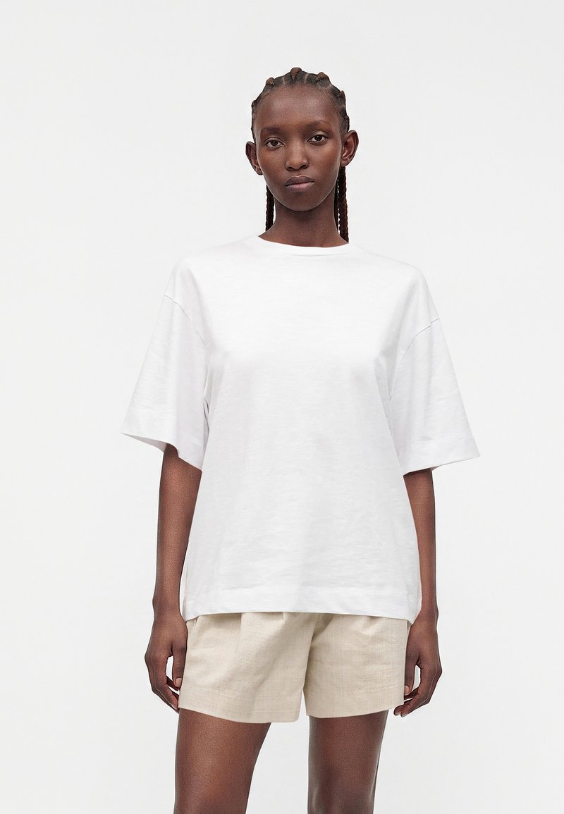 Femme aux cheveux tressés portant un t-shirt blanc uni oversize et un short beige, debout devant un fond blanc.