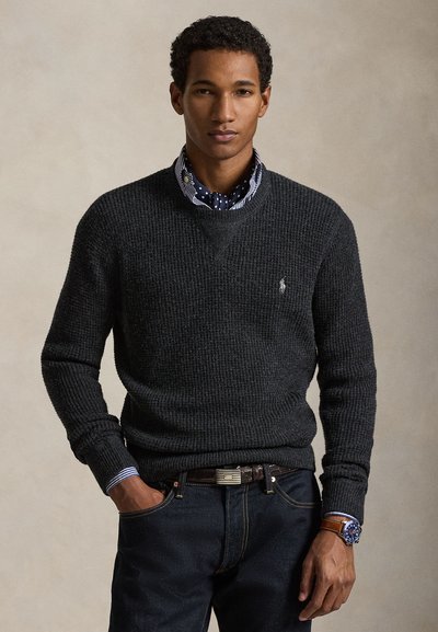 Pull en maille texturée gris foncé avec col en V, porté par-dessus une chemise rayée bleu et blanc avec des pois. Jean en denim foncé et montre.