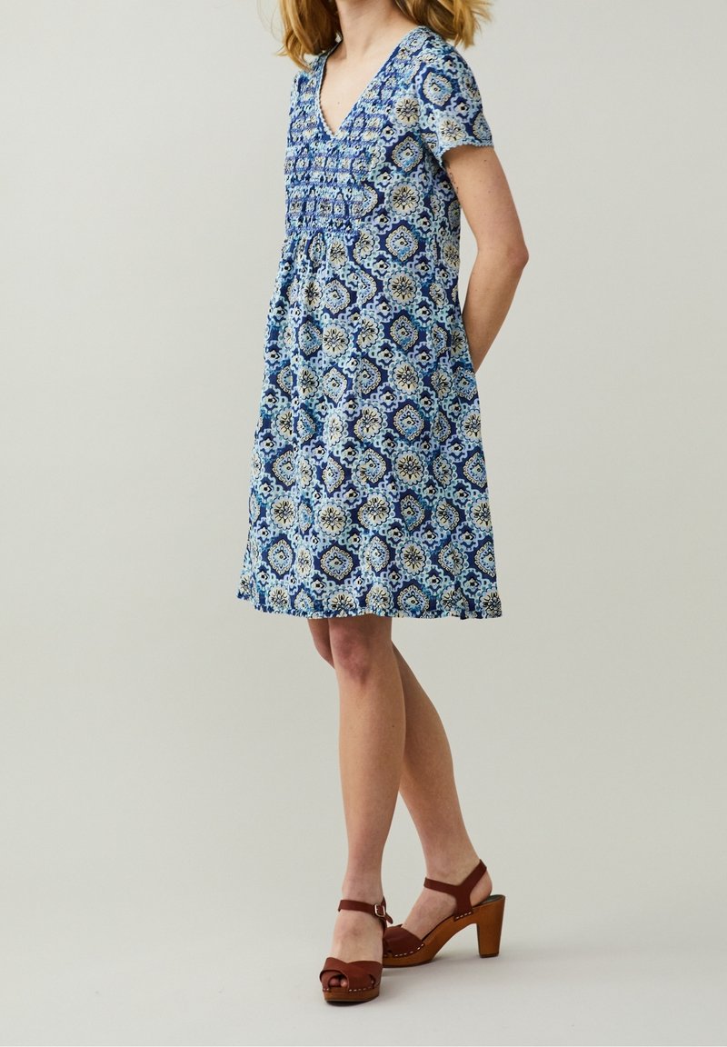 Žena v modro-bílé vzorované šaty do poloviny stehen s krátkými rukávy a hnědými vysokými sandály, stojící na jednofoním pozadí.