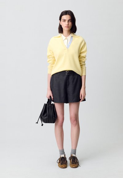Suéter de punto amarillo con collar y botones, combinado con pantalones cortos grises oscuros con detalles de doble hebilla. Sosteniendo una bolsa negra con cordón.
