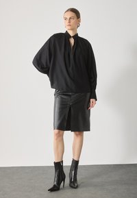 Blusa nera, ampia e svasata con collo alto e spalle arricciate, abbinata a una gonna a tubino in pelle nera con spacco frontale e stivaletti a punta alla caviglia.