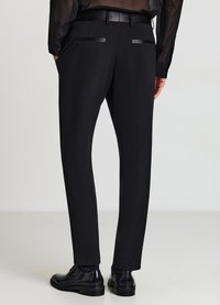 Schwarze, maßgeschneiderte Hose mit elegantem Design aus glattem Stoff, tailliertem Bund und Lederakzenten an den Gesäßtaschen. Kombiniert mit polierten Schuhen.