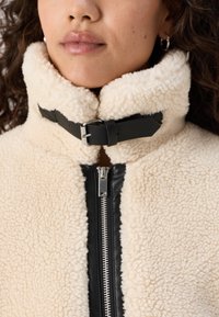 Cream-colored fleece jacket with a black leather zipper and collar, featuring a buckle detail and textured fabric for warmth.