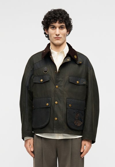 Νεαρός με σγουρά μαλλιά που φοράει σκούρο βαμβακερό μπουφάν με πολλαπλές τσέπες, ορειχάλκινα κουμπιά και καφέ γιακά από κορντόρι.