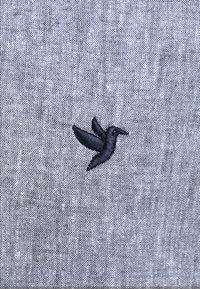 La tela gris presenta un diseño de colibrí bordado en negro en la esquina inferior, mostrando un detallado bordado sobre un fondo texturizado.