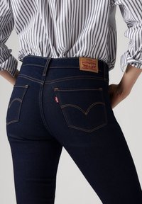 Jean en denim foncé à taille haute, avec une étiquette en cuir beige, des coutures jaunes contrastantes et deux poches arrière aux coutures incurvées.