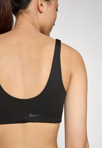 Juodas sportinis liemenukas su apvaliu nugaros iškirpimu, pagamintas iš lygaus audinio, su nedidele Nike logo tamsiame atspaude ant juostos.