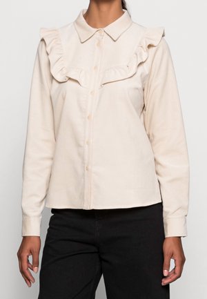 Camisa de botones color crema con detalle de fruncidos en el frente y cuello, confeccionada en tejido texturizado, combinada con pantalones negros.