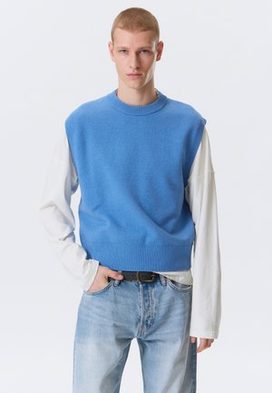 Młody mężczyzna z krótkimi włosami, ubrany w niebieski bezrękawnik na białej koszuli z długim rękawem i jasnoniebieskich dżinsach, stojący z jedną ręką w kieszeni.