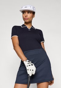 Jūros pajėgų trumpų rankovių polo marškiniai su balta apykakle ir logotipu, derinami su džinsinio audinio tamsiai mėlyna sijonu. Balta golfo pirštinė laiko lazdele; ant galvos balta kepuraitė.