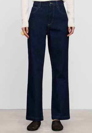 Persoon draagt donkerblauwe jeans met rechte pijpen, wit shirt met lange mouwen, zwarte mesh schoenen en een grote beige ring aan de linkerhand.