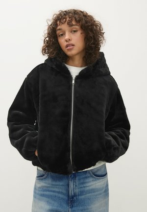 Jeune personne aux cheveux bouclés portant une veste noire en peluche zippée, les mains dans les poches et un jean bleu clair sur un fond uni.