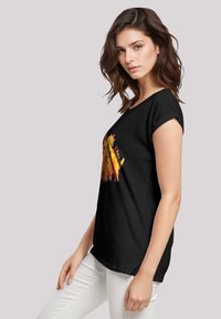 Schwarzes T-Shirt mit kurzen Ärmeln, das ein farbenfrohes Dinosaurier-Motiv zeigt. Der Stoff ist weich und hat eine lässige Passform, kombiniert mit weißen Hosen.