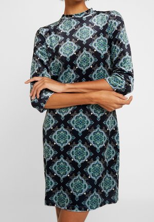 Donna che indossa un vestito di velluto nero con motivi floreali ornate in verde e blu e maniche a tre quarti, con le braccia incrociate in vita.