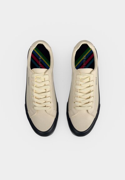 Ζευγάρι μπεζ καμβά sneakers με μαύρες λαστιχένιες μύτες και σόλες, λευκά κορδόνια και πολύχρωμες ριγέ εσωτερικές σόλες, εμφανιζόμενα από πάνω σε λευκό φόντο.