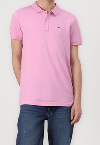 Chemise polo rose en coton, dotée d'un col classique, d'une patte de boutonnage à deux boutons et d'un petit logo sur la poitrine. Portée avec un jean en denim bleu.