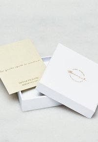 Weißer Pappkarton mit goldgeprägtem Logo, teilweise geöffnet, mit einer beige Karte mit handgeschriebenem Text darin.