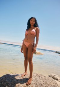 To-delt bikini i blødt lyserødt stof. Toppen har trekantede skåle med flæsekant; underdelen har bindebånd i siderne. Modellen står på en klippe ved vandet.