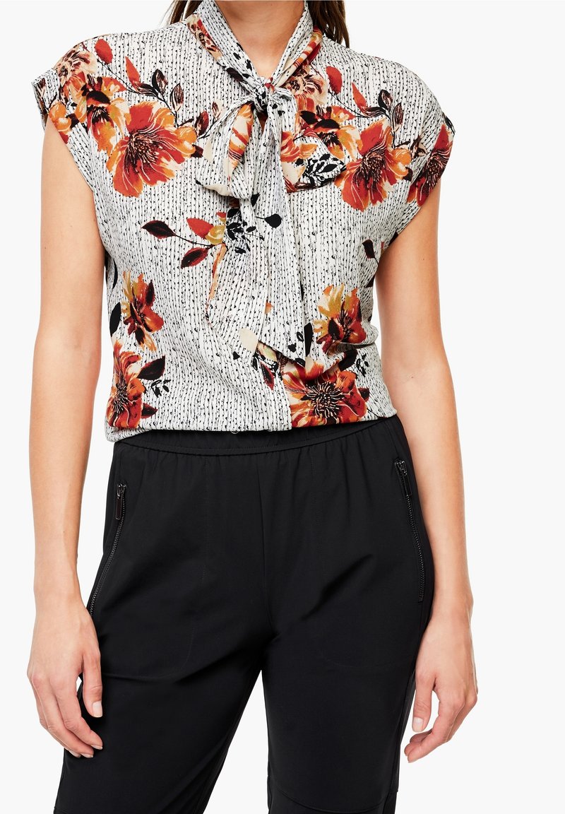 Blusa floreale a maniche corte, con fiori arancioni e neri su uno sfondo bianco strutturato, colletto annodato, abbinata a pantaloni neri.