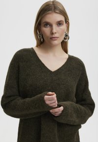 Pulover texturat, fuzos, de un verde închis, cu guler în V și mâneci lungi, având manșete largi și o croială lejeră.