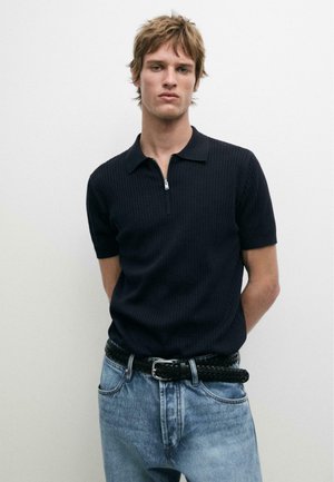 Giovane uomo con capelli scompigliati che indossa una polo nera a maniche corte con zip e jeans azzurri chiari con cintura nera intrecciata, in piedi contro un muro bianco.