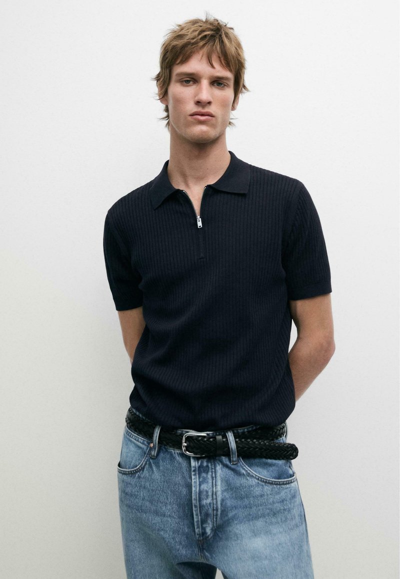 Młody mężczyzna z potarganymi włosami, ubrany w czarną koszulkę polo z krótkim rękawem i suwakiem oraz jasnoniebieskie dżinsy z plecionym czarnym paskiem, stoi na tle białej ściany.