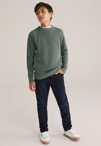 Zaļš teksturēts džemperis ar apaļu kakla izgriezumu un mazu logo, apvienots ar tumši ziliem džinsiem un baltiem sneakeriem ar brūniem zole.