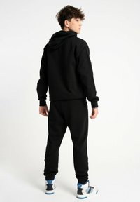 Czarna bawełniana bluza z kapturem na sznurki, połączona z dopasowanymi czarnymi dresami. Posiada ściągacze w mankietach oraz białe i niebieskie sneakersy.