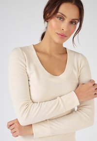 Haut crème à manches longues avec une texture côtelée, décolleté en V profond et manches ajustées, présentant des coutures légères et une finition douce.