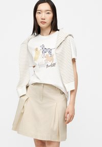 Jeune femme portant un T-shirt blanc à motif de chien, une jupe plissée beige et un pull rayé drapé sur ses épaules.