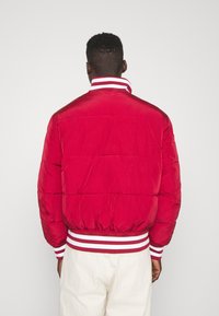 Röd puffjacka med vit och röd randig ribbad krage och muddar. Har quiltad textur och elastisk nederkant. Bakvy visas.