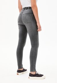 Personne portant un jean skinny gris taille haute et des baskets basses noires avec des semelles blanches, debout devant un fond blanc.